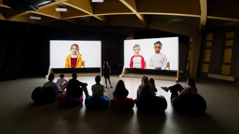 People in a dark exhibition space silhouetted against two screens showing a projection of a film of children talking
